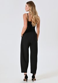 Schwarzer Jumpsuit mit dünnen Trägern, einem taillierten Oberteil und weiten Hosenbeinen. Glattes Material und seitliche Taschen. Kombiniert mit schwarzen High Heels.