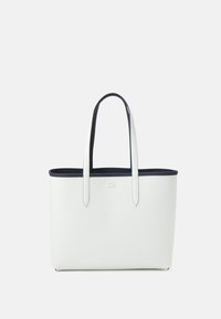 Sac fourre-tout blanc texturé avec une bordure et des anses bleu marine, présentant un petit logo sur le devant. Design élégant avec un dessus ouvert.