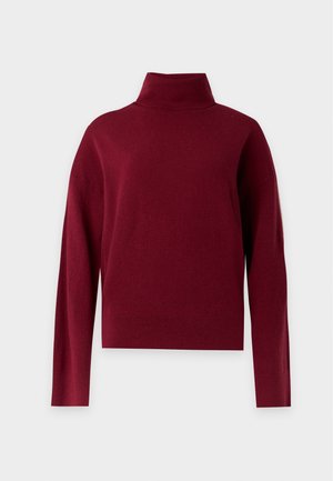 Femme portant un pull col roulé bordeaux, une jupe beige avec un motif en dentelle florale noire et des bottines noires, debout devant un fond blanc.