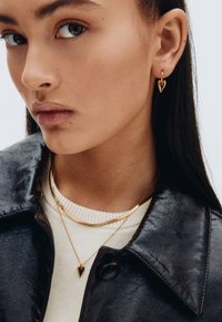 Goldene, herzförmige Ohrringe und gestapelte goldene Halsketten. Das Model trägt eine schwarze Lederjacke und einen weißen, gerippten Pullover. Glatte, glänzende Texturen.