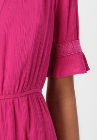 Fuchsia jurk van een textuur, geplisseerde stof. Heeft een ceintuur in de taille en decoratieve kantafwerking op de mouwen. Korte fluttermouwen.