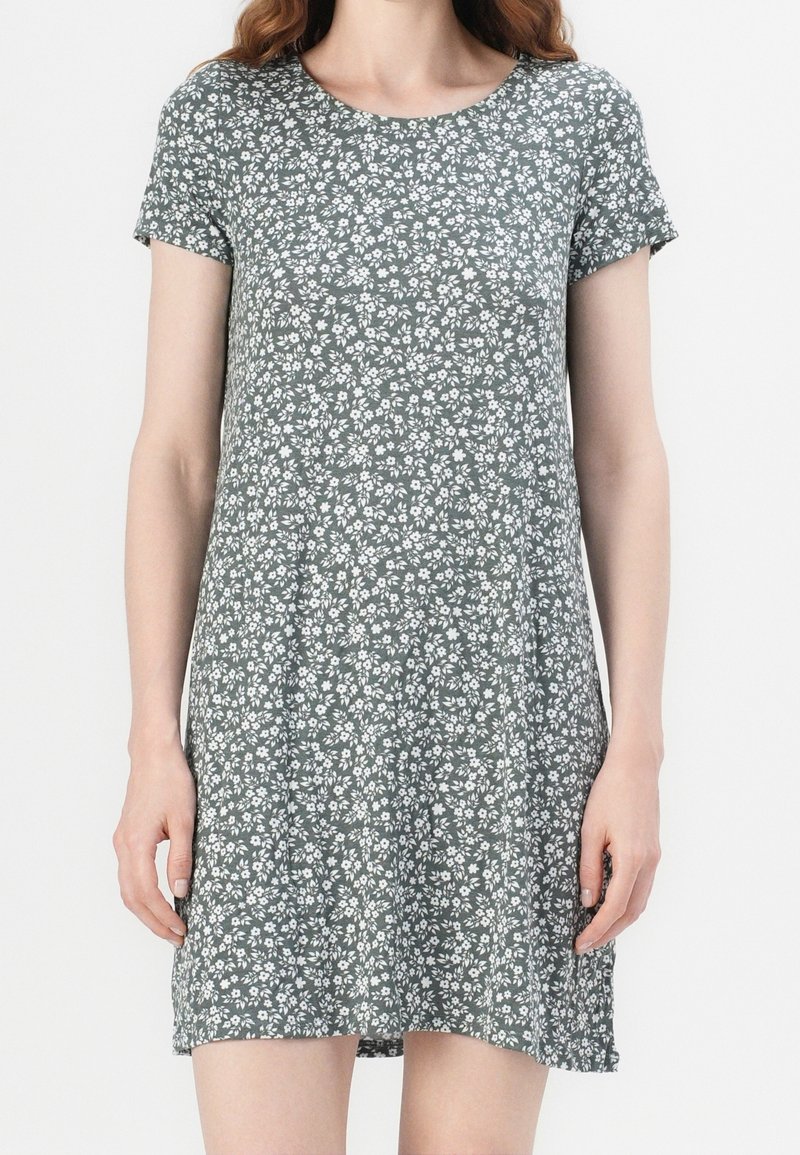 Femme portant une robe verte à manches courtes et longueur au genou avec un motif floral blanc, debout devant un fond uni.