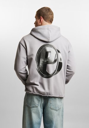 Mężczyzna w jasnoszarej bluzie z dużą metaliczną literą 3D „P” na plecach, w połączeniu z jasnoniebieskimi dżinsami, na jednolitym tle.