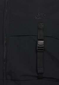 Zwarte Nike-jack met een rits aan de voorkant, geweven stofstructuur, verstelbare band en gespdetail, en reliëflogo op de borst.