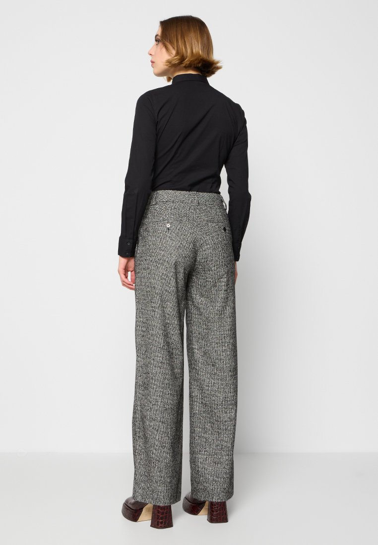 Frau in einem schwarzen Langarmoberteil, graue gemusterte weitgeschnittene Hose und braune High Heels, die mit dem Gesicht zu einer weißen Wand steht.