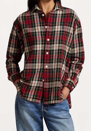 Person trägt ein rot-grün-weiß kariertes Hemd mit langen Ärmeln und blaue Jeans, Hände in den Taschen.