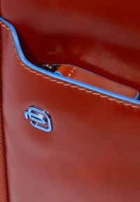 Bolso de cuero marrón con una textura suave, que presenta detalles en azul y un logotipo azul prominente. Incluye un bolsillo interior con un llavero.