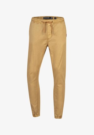 Man draagt beige joggerbroek met elastische boorden en een bruine trekkoord, gecombineerd met zwarte Adidas sneakers met witte zolen.