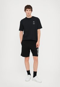 Svart T-skjorte og svarte shorts antrekk. T-skjorten har et lite trykt logo; shortsene har en kontrasterende hvit grafikk på ett ben. Hvite joggesko.
