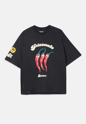 T-shirt nera oversize in cotone, con grafiche di peperoncini rossi contornati di verde e il testo "Piccante Barrow" in crema.