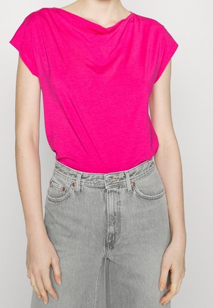 Femme portant un haut rose vif à manches courtes rentré dans un jean gris clair taille haute, sur un fond blanc uni.