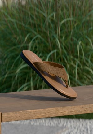 Brauner Riemchensandalette mit strukturiertem Riemen und schwarzer Sohle, balanciert am Rand einer Holzbank, grüne Pflanzen im Hintergrund unscharf.