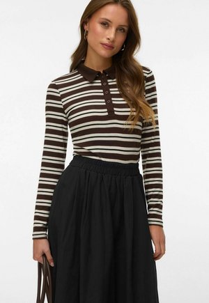 Pikkade juustega naine, kandes kuldsete rõngastena kõrvarõngaid, pruunide ja valgete triipudega pika käisega polosärgiga, musta seelikut ja hoides käes pruuni koti sangasid.