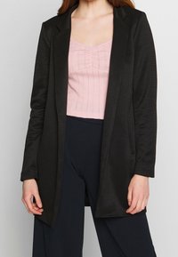 Blazer noir sur mesure avec un design uni, une texture lisse et un col cranté. Il possède des poches latérales et un devant ouvert. Porté sur un haut rose.