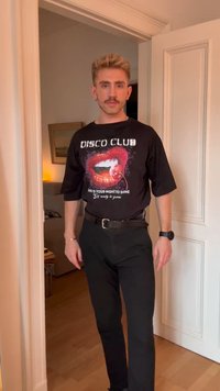 Muž s fúzami, oblečený v čiernom tričku "Disco Club" s grafikou pier, stojí v dverách v teplo osvetlenej miestnosti.