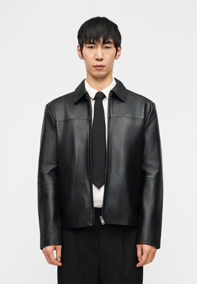 Homme portant une veste en cuir noire par-dessus une chemise blanche et une cravate noire, debout devant un fond clair uni, faisant face à l'avant.