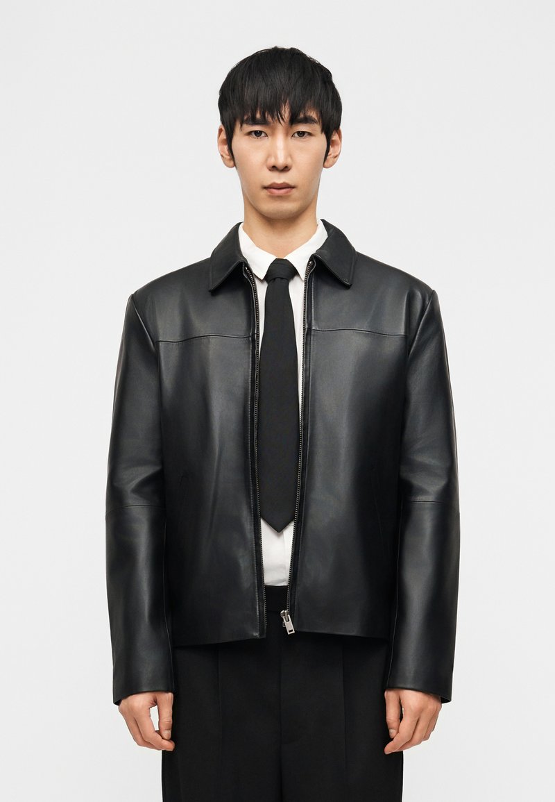 Άνδρας που φοράει μαύρο δερμάτινο μπουφάν πάνω από λευκό πουκάμισο και μαύρη γραβάτα, στέκεται μπροστά σε ένα απλό ανοιχτόχρωμο φόντο, κοιτώντας προς τα εμπρός.