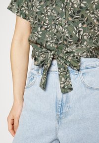Camisetta verde a maniche corte con stampa di foglie, legata in vita, abbinata a pantaloni corti di jeans azzurri a vita alta.
