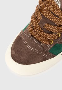 Sneaker in suede marrone con accenti verdi, lacci marroni texturizzati e suola in gomma bianca. Presenta dettagli cuciti attorno alla punta.