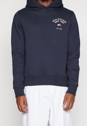 Man wearing navy blue Tommy Hilfiger hoodie with white text and logo, paired with loose white pants, standing against plain background.