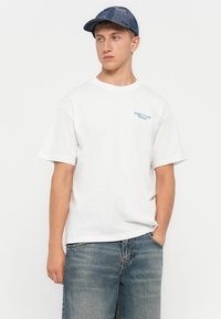 Hvit bomulls T-skjorte med korte ermer og en blå logo på venstre bryst, brukt sammen med falmede blå denimshorts og en mønstret blå caps.