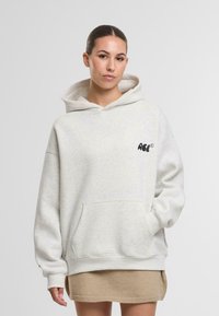 Felpa grigia chiara in tessuto morbido con tasca frontale, dotata di un logo nero sul lato sinistro del petto e polsini a costine. Abbinata a una gonna beige.