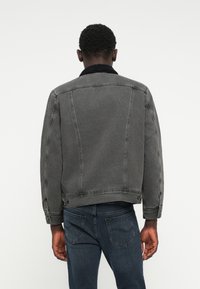 Jachetă din denim de culoare gri închis, cu un material texturat; are un guler negru și pufos și cusături vizibile pe spate.