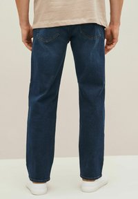 Donkerblauwe spijkerbroek met een recht gesneden model, voorzien van achterzakken, subtiele stikseldetails, en gedragen met witte sneakers.