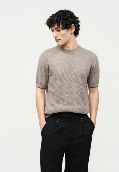 Νέος άνδρας με σγουρά μαλλιά, φορώντας μπεζ υφασμάτινο κοντομάνικο πουλόβερ και μαύρο παντελόνι, με τα χέρια στις τσέπες, κοιτάζει προς τα κάτω και πλάγια.