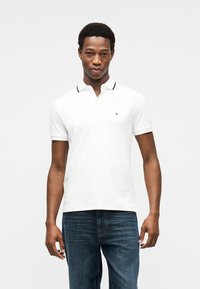 Λευκό πόλο μπλουζάκι με αντίθετο σκούρο μπλε γιακά, κοντά μανίκια και μικρό λογότυπο στο στήθος· συνδυάζεται με σκούρο μπλε τζιν.