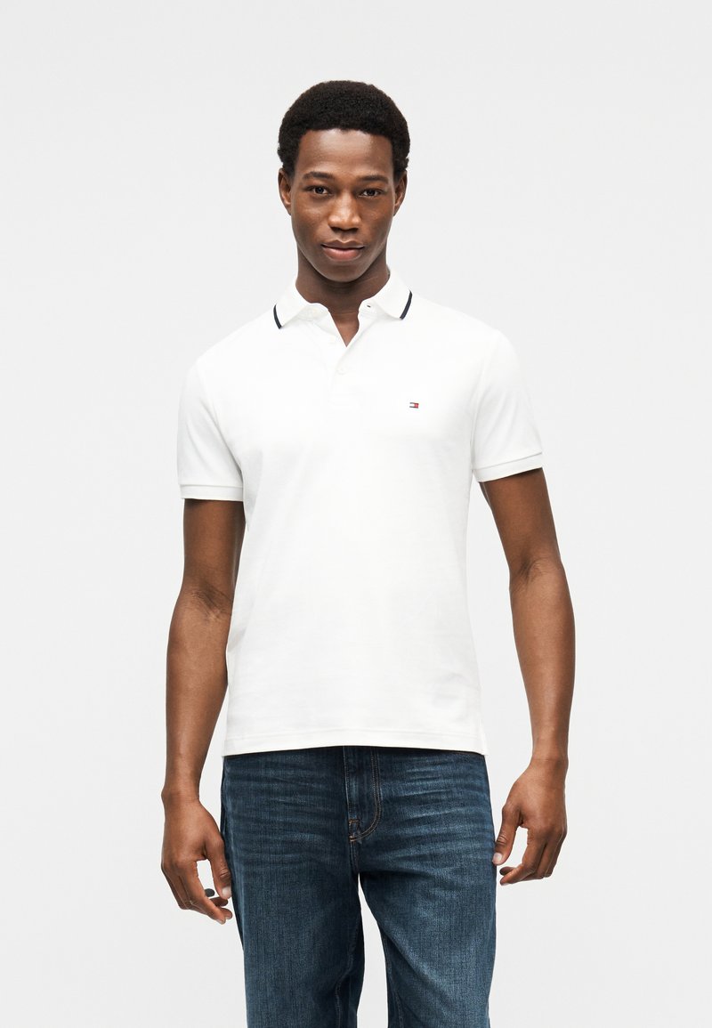 Λευκό πόλο μπλουζάκι με αντίθετο σκούρο μπλε γιακά, κοντά μανίκια και μικρό λογότυπο στο στήθος· συνδυάζεται με σκούρο μπλε τζιν.