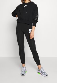 Sudadera corta negra con logo, combinada con leggings negros que cuentan con un gráfico blanco de "J". Zapatillas grises con detalles verdes completan el look.