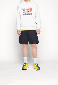 Felpa grigia chiaro con una grafica rossa e nera, t-shirt gialla sotto, pantaloni corti blu navy, calzini bianchi e scarpe da ginnastica multicolori.
