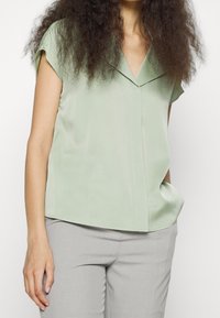Ljusgrön, kortärmad blus med V-ringning, tillverkad av mjukt tyg. Har en avslappnad passform och rundad nederkant. Bärs med grå byxor.