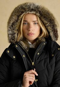 Blondide juustega naine, kes kannab musta kasukat, mille kapuuts on karvase vooderdusega, hoiab tõmblukkude juures kaela ja vaatab otse ette beez taustal.