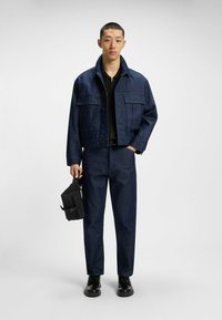 Giacca di jeans e pantaloni di un blu scuro, con una maglietta nera con zip sotto. Il modello tiene una borsa nera con una fibbia. Scarpe nere completano il look.