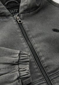 Sudadera con cremallera gris de textura suave, con cremallera negra y puños acanalados. Incluye un logo en el pecho y una capucha.