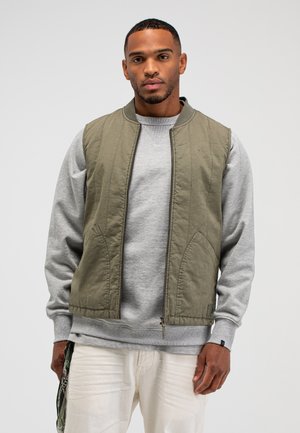 Mann trägt olivgrüne Weste mit Reißverschluss über grauem Sweatshirt und weißer Hose, steht vor einfachem hellem Hintergrund.