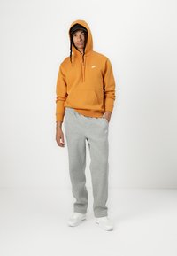 Sweatshirt laranja com capuz e bolsa frontal, calças de moletom cinzentas e ténis brancos. Textura suave, corte casual e detalhes com o logo da Nike.