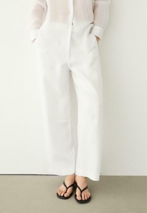 Osoba stojąca, ubrana w luźne białe lniane spodnie, biały przezroczysty koszulowy top na guziki oraz czarne japonki, na neutralnej podłodze.