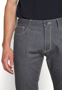 Man i mörkgrå jeans med vit söm och en svart tröja, står med vänsterhanden avslappnad vid sidan.