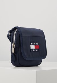 Borsa a tracolla in nylon blu navy con chiusura a pattina, zip argentata e patch logo ricamato con accenti in rosso, bianco e blu. Design compatto e rettangolare.