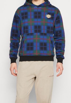 Mann trägt einen blau-grün-rot karierten Hoodie mit schwarzen Bündchen und beige Hose mit leichter Stickerei am linken Oberschenkel.