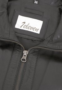 Innenansicht einer schwarzen Lederjacke mit einem YKK-Reißverschluss, ausgestattet mit einem Etikett, auf dem "Televen" steht, und einem Größenschild mit der Größe "36". Glatte Textur.