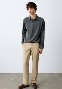 Tânăr purtând un tricou polo cu mâneci lungi gri închis, pantaloni bej și adidași maro, stând în picioare în fața unui perete simplu și deschis la culoare.