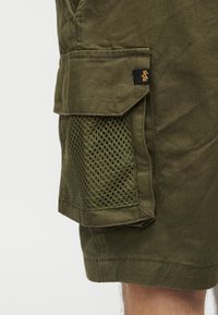 Χακί σορτς με δίχτυ τσέπης cargo και μια μικρή μαύρη και κίτρινη υφασμάτινη ετικέτα στο καπάκι της τσέπης.