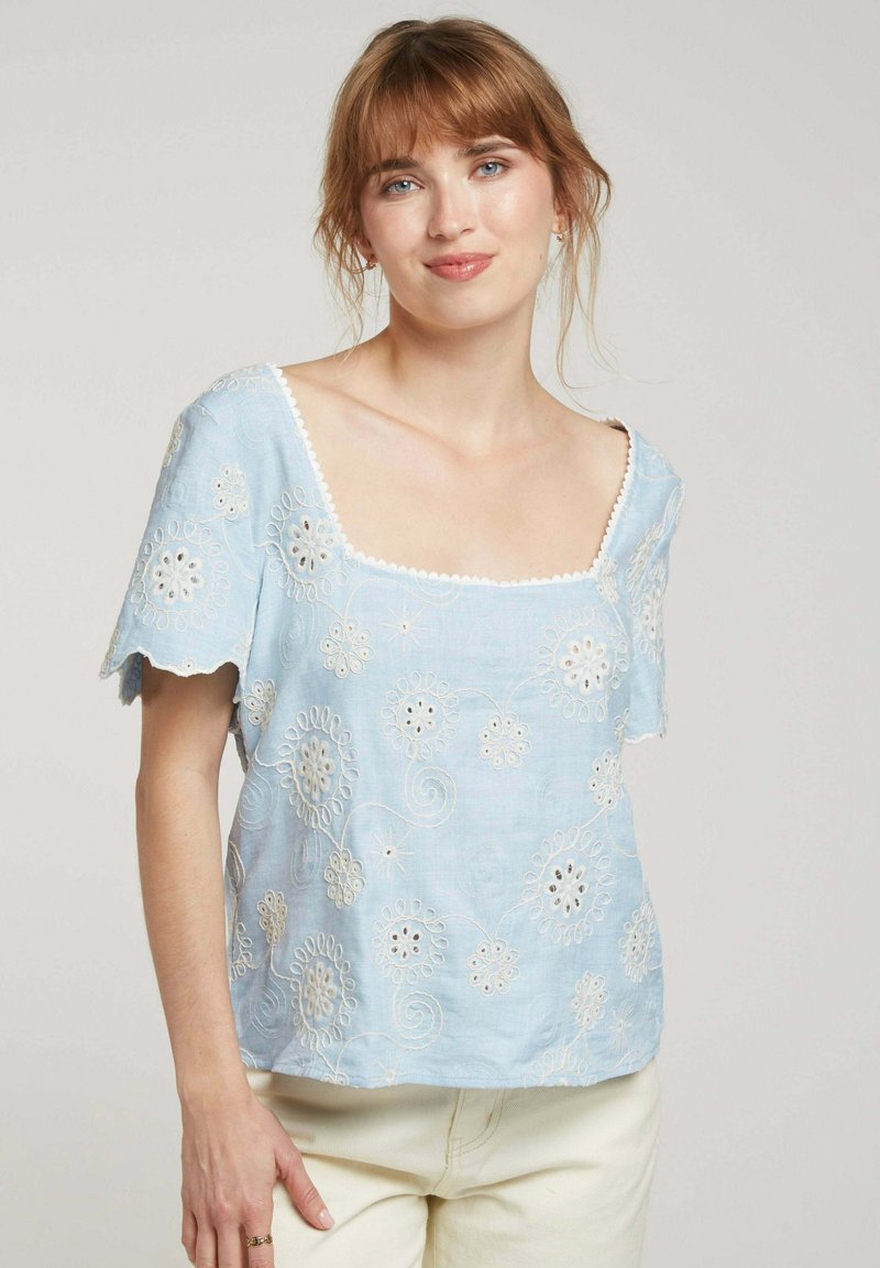 Blouse brodée bleu clair à manches courtes, décolleté carré, motifs floraux détaillés et bords festonnés. Texture semblable à du lin.