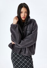 Veste en polaire grise avec une surface douce et texturée, dotée d'une poche zippée et d'une fermeture à pression. Associée à une jupe noire à motifs.