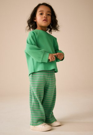 Lille barn med krøllet hår iført en grøn ribbet sweater og matchende stribede bukser med hvide sneakers, stående på en neutral baggrund.