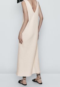 Langes, ärmelloses Kleid aus strukturiertem beigem Stoff mit einem tiefen V-Ausschnitt am Rücken. Kombiniert mit schwarzen Riemchensandalen. Insgesamt minimalistisches Design.